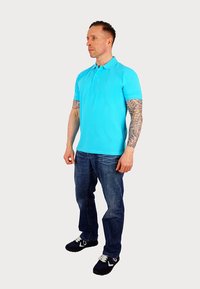 Turkos polo shirt med korta ärmar, matchad med blåjeans och mörka sneakers. Märkbara tatueringar på armarna.