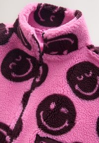 Veste en polaire rose avec motifs de visages souriants noirs et fermeture éclair à l'avant, présentée pliée avec le col relevé.