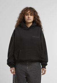 Schwarzer Kapuzenpullover mit einer Fronttasche und dem Wort "VERBOTEN" in einem kontrastierenden Grau. Verfügt über überschnittene Schultern und eine lockere Passform.