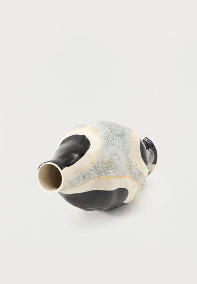 Μικρό κεραμικό βάζο με ακανόνιστα μαύρα και υφασμένα ανοιχτό μπλε-γκρι μοτίβα σε λευκή βάση, οριζόντια τοποθετημένο σε λευκή επιφάνεια.