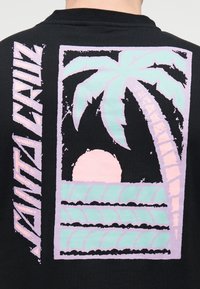 Μαύρο πίσω μέρος μπλούζας με γραφικό απαλό ροζ και μέντα που απεικονίζει φοινικόδεντρο, ήλιο που δύει πάνω από τα κύματα του ωκεανού και το κείμενο "VANPALAZ" στην αριστερή πλευρά.