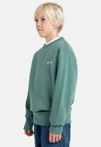 Felpa verde con colletto a girocollo e polsini a costine, con un piccolo logo beige sul petto. Abbinata a jeans in denim blu.