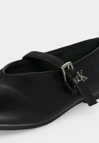 Chaussure plate en cuir noir avec une fine bride et une boucle argentée affichant un petit logo "ck" sur le côté.