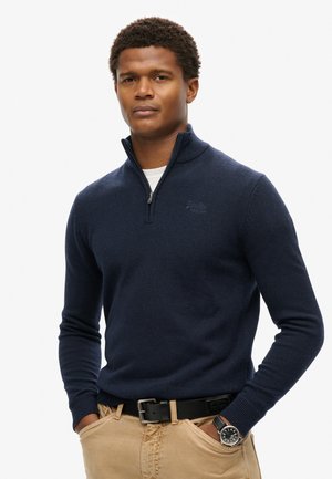 Marineblauwe zip-up trui met een hoge kraag, geribbelde manchetten en een subtiele logo op de borst, gecombineerd met beige broek en een zwarte horloge.