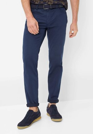 Mann trägt marineblaue, schmal geschnittene Hose, schwarzen geflochtenen Gürtel und dunkelblaue Wildledersneaker mit Gummisohlen, steht vor einem weißen Hintergrund.