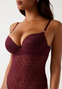 Body color borgogna con sovrapposizione in pizzo, dotato di coppe con ferretto, spalline regolabili e motivi tonali intricati. Texture liscia e trasparente.