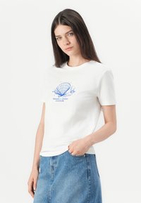 Hvid bomulds T-shirt med blåt grafik af en muslingeskal og teksten "SEASHELL SEEKER SINCE 1996." Korte ærmer og rund halsudskæring.
