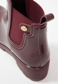 Bottine en caoutchouc bordeaux avec un panneau élastique texturé, un design courbé et une petite boucle à l'arrière ; étiquette dorée accrochée.