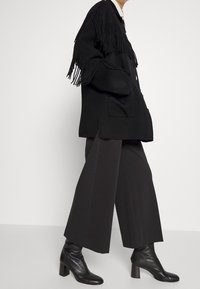 Zwarte oversized cardigan met franjes op de schouders, voorzakken en knoopsluiting, gecombineerd met zwarte brede broekspijpen en hoge laarzen.