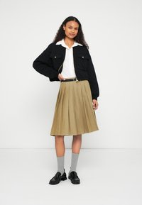 Μαύρο καπιτονέ μπουφάν με δύο τσέπες στο στήθος, λευκό πουκάμισο με γιακά, πτυχωτή χακί φούστα, γκρι κάλτσες και μαύρα μοκασίνια.