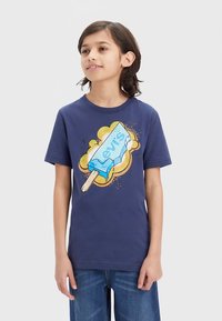 Niño con camiseta azul marino con un gran gráfico de una barra de helado azul de Levi's y salpicadura amarilla, combinado con vaqueros azules.