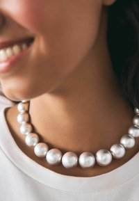 Srebrna ogrlica z okroglimi, sijočimi biseri različnih velikosti, ki počiva na ključnici preko enostavnega belega vrha.