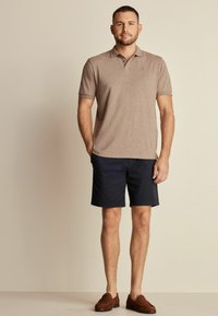 Polo de color marrón con mangas cortas, combinado con pantalones cortos azul marino y mocasines marrones. Textura suave con un sutil logo en la camiseta.