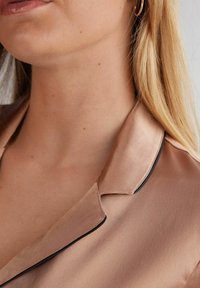 Satinbluskrage i mjuk beige med svarta kanter, visar en slät textur, subtil lyster och öppet halslinjedesign.