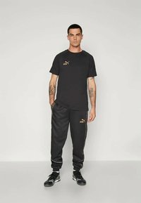 Vīrietis ar tetovējumiem, ģērbies melnā Puma King t-kreklā un sporta biksēs, stāv ar vienu roku kabatā uz vientuļa gaiša fona.