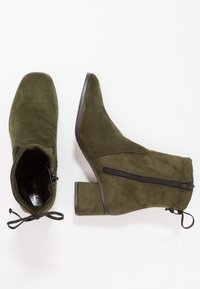 Botas de tobillo de ante verde oliva con punta afilada, tacón cuadrado, cierre de cremallera lateral y detalle de cordones decorativos en la parte trasera.