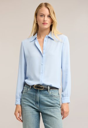 Kvinde iført en lyseblå knappet bluse stoppet ned i lyseblå jeans med et sort bælte, der står mod en ensfarvet beige baggrund.