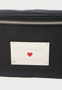 Juodas tekstūruotas maišas su sidabriniu užtrauktuku ir sidabrine etikete, ant kurios yra užrašas "LOVE MOSCHINO", raudona širdies simbolika ir įrašas "Pagaminta su meile".