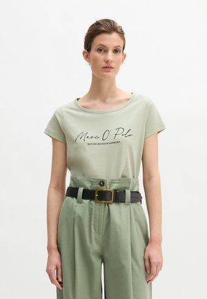 Kobieta ubrana w jasnorozowy T-shirt marki Marc O' Polo z napisem "Nature Moves in Harmony", zielone spodnie z wysokim stanem oraz czarny pasek ze złotą klamrą.