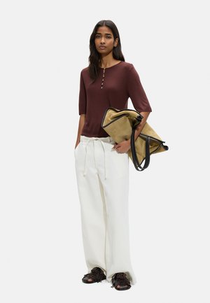 Kobieta ubrana w brązową bluzkę zapinaną na guziki, białe szerokie spodnie, czarne sandały z otwartymi palcami, trzymająca dużą beżowo-czarną torbę na ramię.