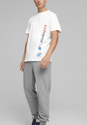 Homme portant un t-shirt BMW Motorsport blanc avec texte et logo rouges et bleus, associé à un pantalon de survêtement gris sur un fond gris uni.