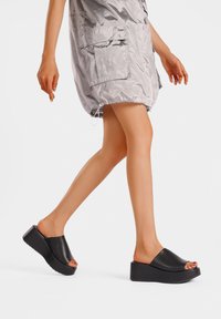 Sandali piattaforma neri con design open toe, texture in pelle liscia e suola spessa, abbinati a un vestito lucido grigio chiaro.