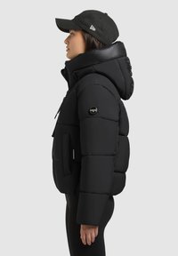 Juodas pūkuotas švarkas su aukštu apykakle ir kapišonu, su priekinėmis kišenėmis ir tekstūruota medžiaga. Priedas - juoda kepurė.