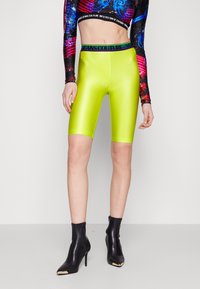 Neonově zelené cyklistické kraťasy s hladkým, lesklým povrchem a černým páskem se značkovým logem, v kombinaci s černými špičatými kotníkovými botami.