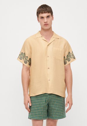 Beige Kurzarmhemd mit grüner Spiralstickerei an den Ärmeln, leichtes Material, einfacher Kragen und einer einzelnen Brusttasche.