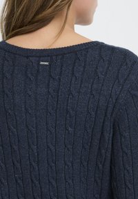 Námornícky modrý sveter s pleteným vzorom, ktorý má široký výstrih, rebrované manžety a malú nálepku na zadnej strane pri golieri. Mäkká textúra.