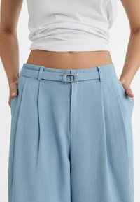 Lichtblauwe high-waisted broek met een riem met een rechthoekige zilveren gesp, geplooide voorkant en zijzakken. Soepele stof textuur.
