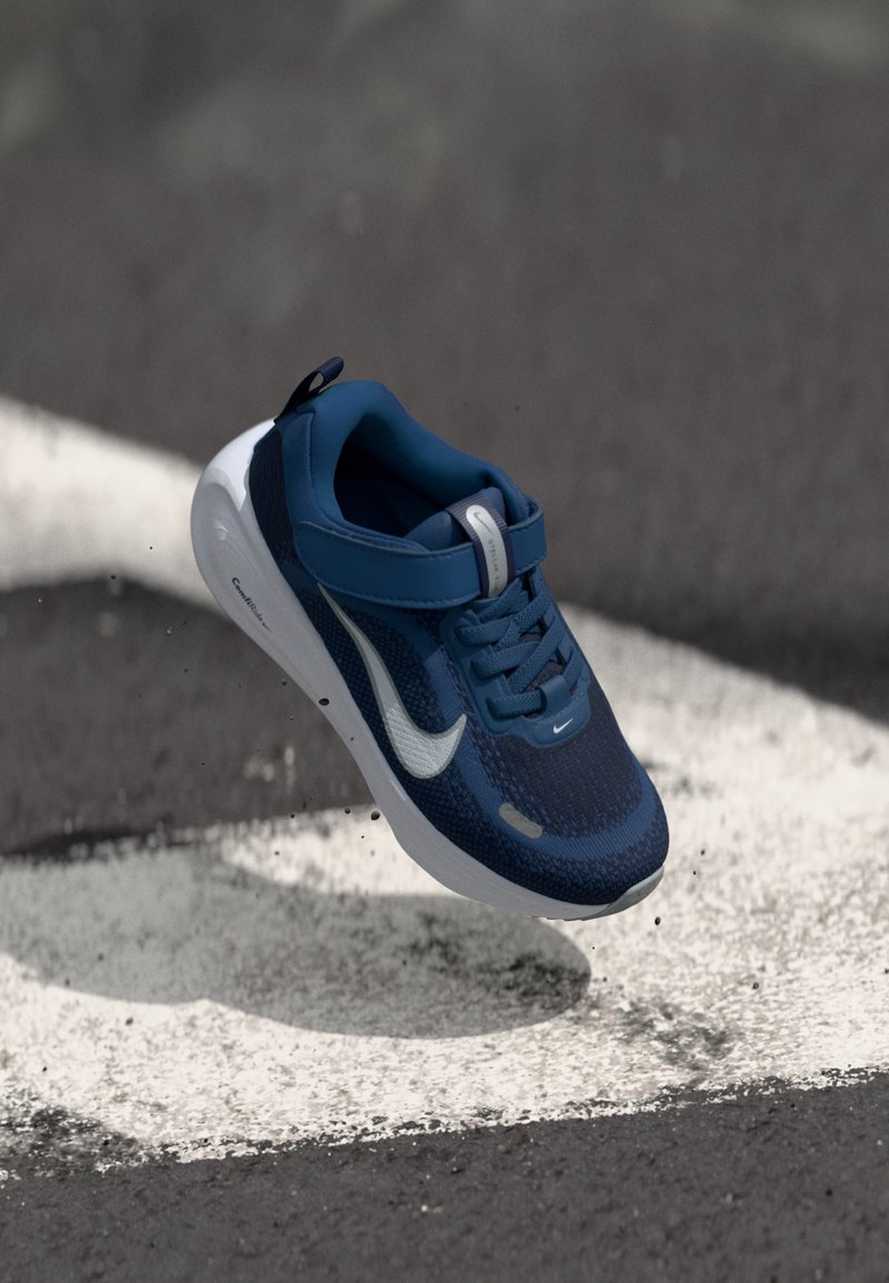 Mėlynas sportinis batelis su tekstūruota tinklelio viršutine dalimi, baltomis detalėmis, lygiu padu ir reguliuojama juosta; sukurtas komfortui ir palaikymui.