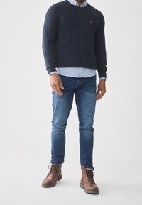 Marineblauer, gestrickter Pullover mit strukturiertem Zopf-Muster, rotem Logo auf der Brust, über einem hellblauen Hemd getragen, kombiniert mit blauen Jeans und braunen Schnürstiefeln.