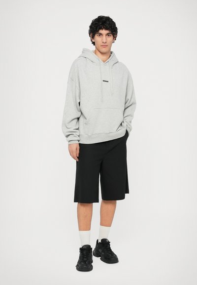 Felpa grigia con cappuccio e tasca a marsupio, abbinata a pantaloni corti neri fino al ginocchio e sneakers nere con motivo testurizzato.