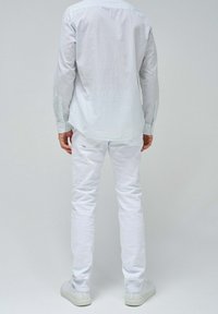 Camicia bianca a maniche lunghe con righe verticali blu chiaro, abbinata a pantaloni bianchi slim-fit e sneakers bianche, vista da dietro.