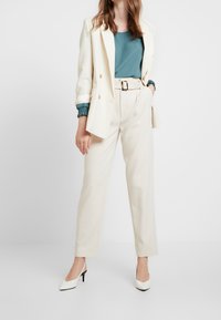 Blazer crème porté sur un chemisier bleu sarcelle, associé à un pantalon beige taille haute et des escarpins blancs à bouts pointus. Caractérisé par une coupe ajustée et une ceinture.