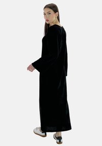 Vestido largo de terciopelo negro con puños acampanados, con un escote redondeado. La modelo lleva zapatillas blancas con detalles en negro.