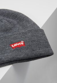 Grå strikket hue med en tætsiddende pasform, der har et rødt Levi's logo patch. Teksturen er blød og præsenterer et enkelt, klassisk design.