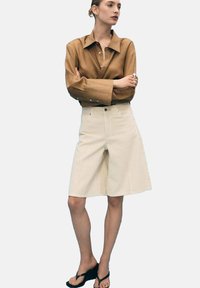 Chemise à manches longues marron avec patte de boutonnage, associée à un short crème à mi-cuisses. Les chaussures sont des tongs noires. Design simple et couleurs neutres.