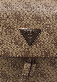 Borsa a tracolla marrone con motivo e finitura testurizzata. Presenta un logo triangolare Guess nero, con dettagli marroni e una chiusura in metallo.