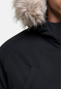 Cappotto nero con un morbido bordo in pelliccia sintetica di colore marrone chiaro. Il tessuto appare liscio con una finitura opaca e presenta un dettaglio con tasche laterali.