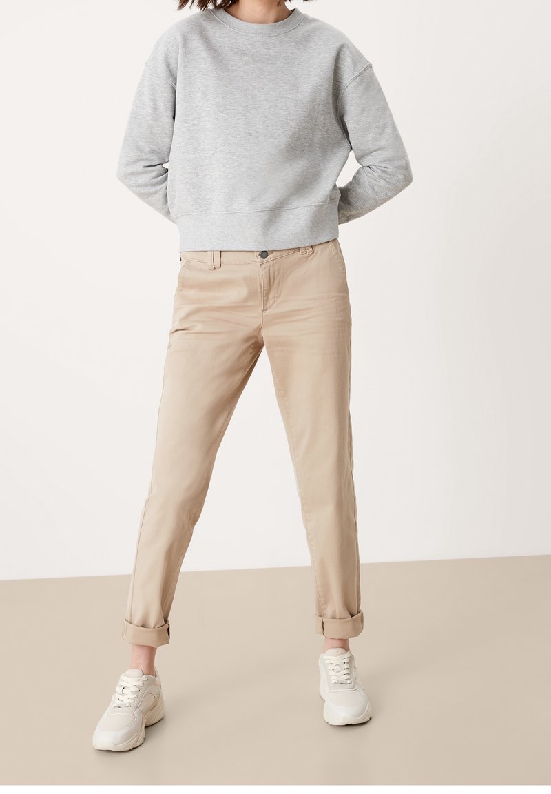 Sweat-shirt gris avec un col rond et des manches longues, associé à un chino beige retroussé et des baskets blanches. Tenue décontractée sur un fond neutre.