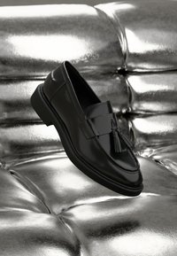 Mocassin en cuir noir avec détail pompon, semelle plate et texture lisse, présenté au-dessus d'une surface métallique brillante et capitonnée.
