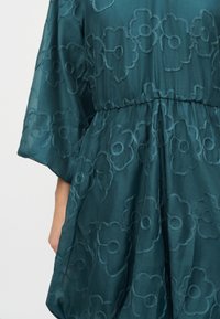 Τιρκουάζ φόρεμα με σφηκωμένη μέση και μεγάλο κεντητό λουλούδι που καλύπτει το διάφανο ύφασμα, εμφανιζόμενο στον κορμό και το χέρι ενός ατόμου.