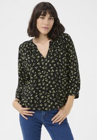 Blouse noire avec imprimé floral jaune, en V, manches 3/4, détails froncés aux épaules, fabriquée en tissu léger. Assortie avec un jean bleu.