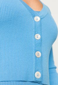 Lyseblå ribbestrikket cardigan med V-hals og fire store hvite skjellknapper foran. Myk tekstur med figurvennlig fasong.