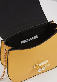 Bolso de mano de imitación de cuero amarillo con un cierre y cadena en tono dorado. Interior forrado en tela negra; la etiqueta dice "PIECES". Textura suave.