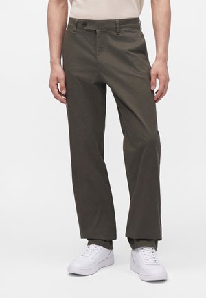 Personne portant un pantalon droit vert olive et des baskets blanches, debout devant un fond clair uni.
