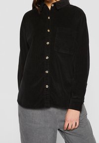 Chemise boutonnée en velours côtelé noir avec col, poche avant et boutons beiges ; présente un tissu texturé et des manches longues.