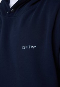 Sweat à capuche bleu marine avec une texture douce, arborant un logo brodé blanc "ESTEEM" sur la poitrine gauche, et des accents à boutons-pression sur le col.